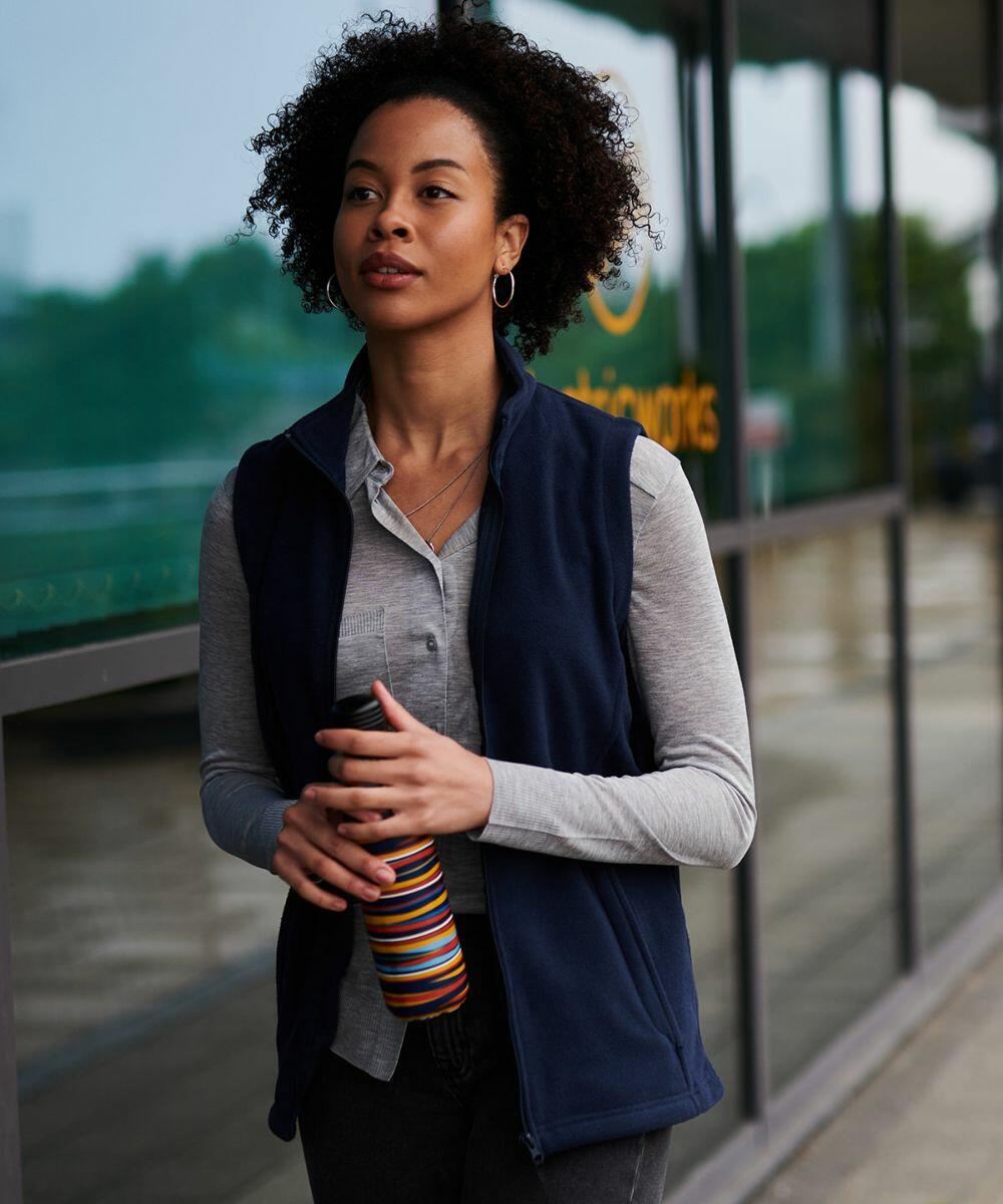 A person with curly hair wearing a navy vest and gray shirt holds a colorful striped RG186 by Regatta while standing outdoors near a windowed building.