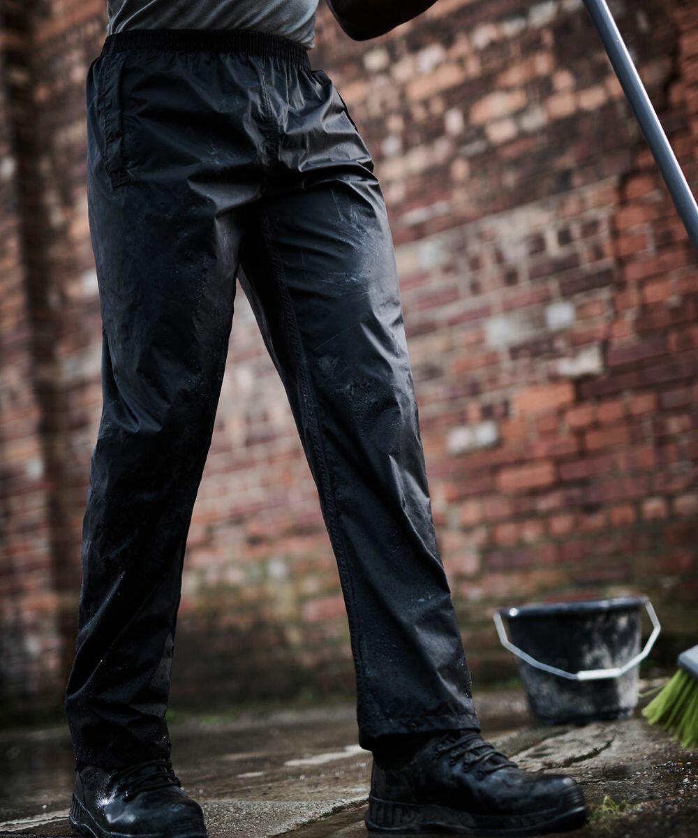 Person wearing black Regatta RG208 waterproof pants standing on wet ground near a brick wall, holding a broom. There is a bucket filled with water in the background.