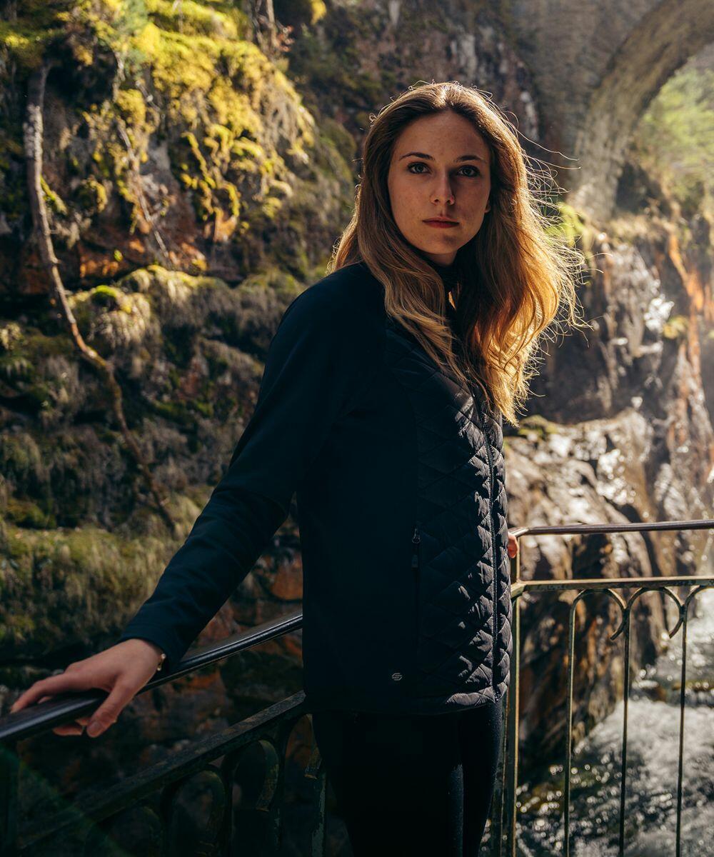 A person with long hair stands outdoors on a metal railing, surrounded by moss-covered rocks and sunlight, wearing the ST196 jacket from Stormtech.