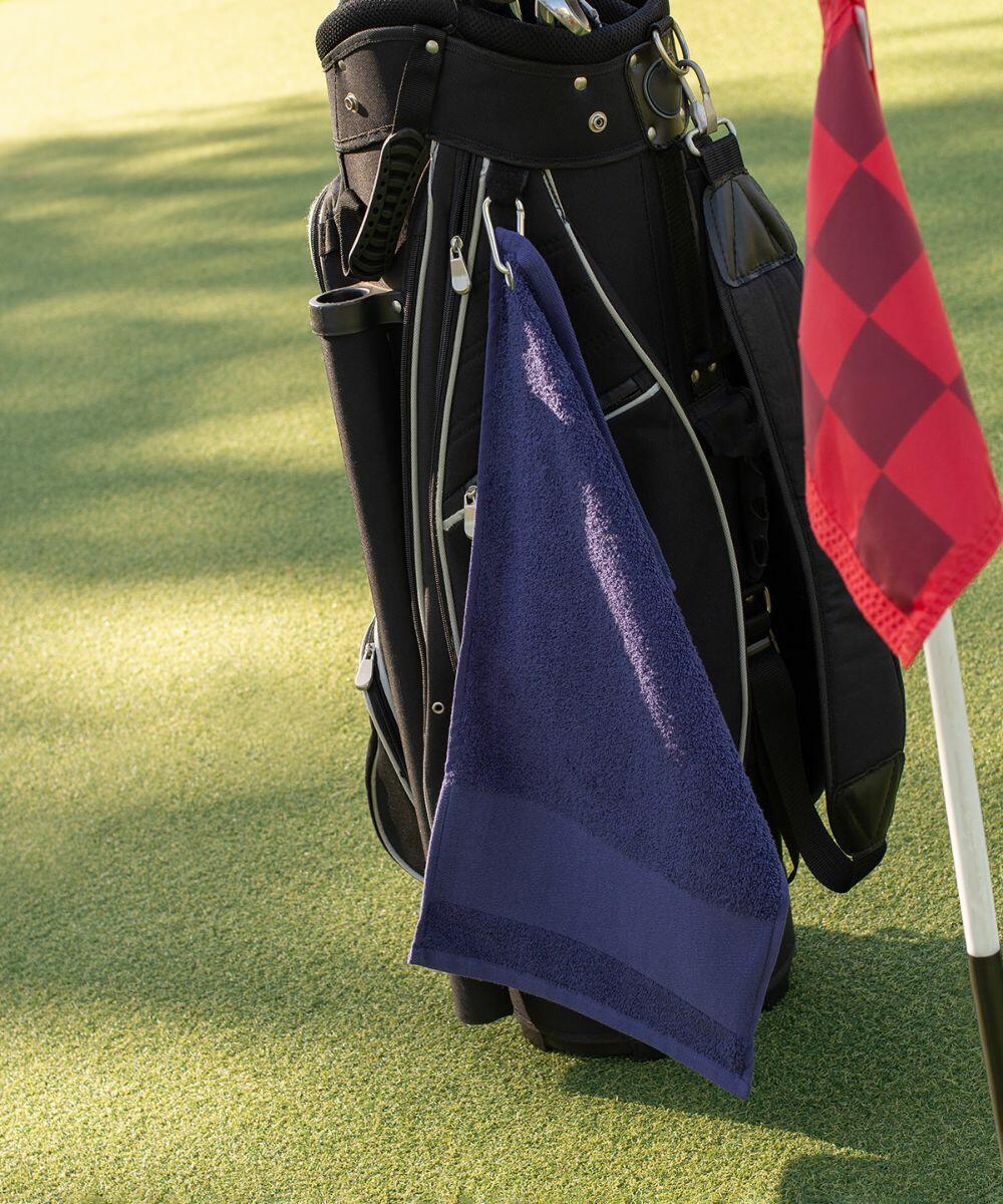 A black golf bag with a blue Towel City TC033 hanging from it is placed on a green golf course, next to a red and black checkered flag.