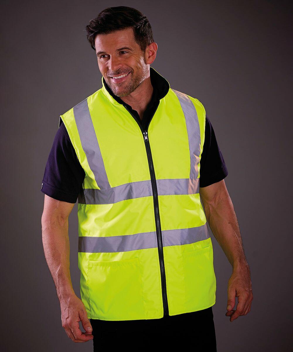 A man wearing a Yoko YK041 high-visibility yellow safety vest with grey reflective stripes stands and smiles.