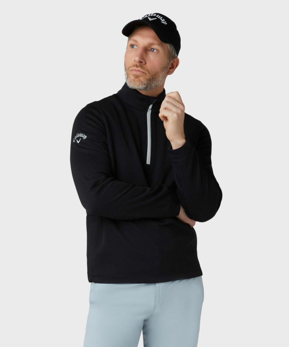 A man wearing a black pullover and light grey pants stands against a plain background. He is also wearing a Callaway CW089 with a white logo. His right hand is raised near his face.
