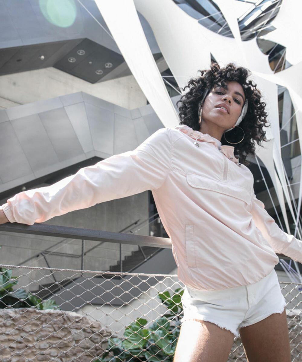 A person wearing a light pink jacket and white shorts poses in front of a modern architectural structure with white fabric installations, sporting the BY095 jacket by Build Your Brand.