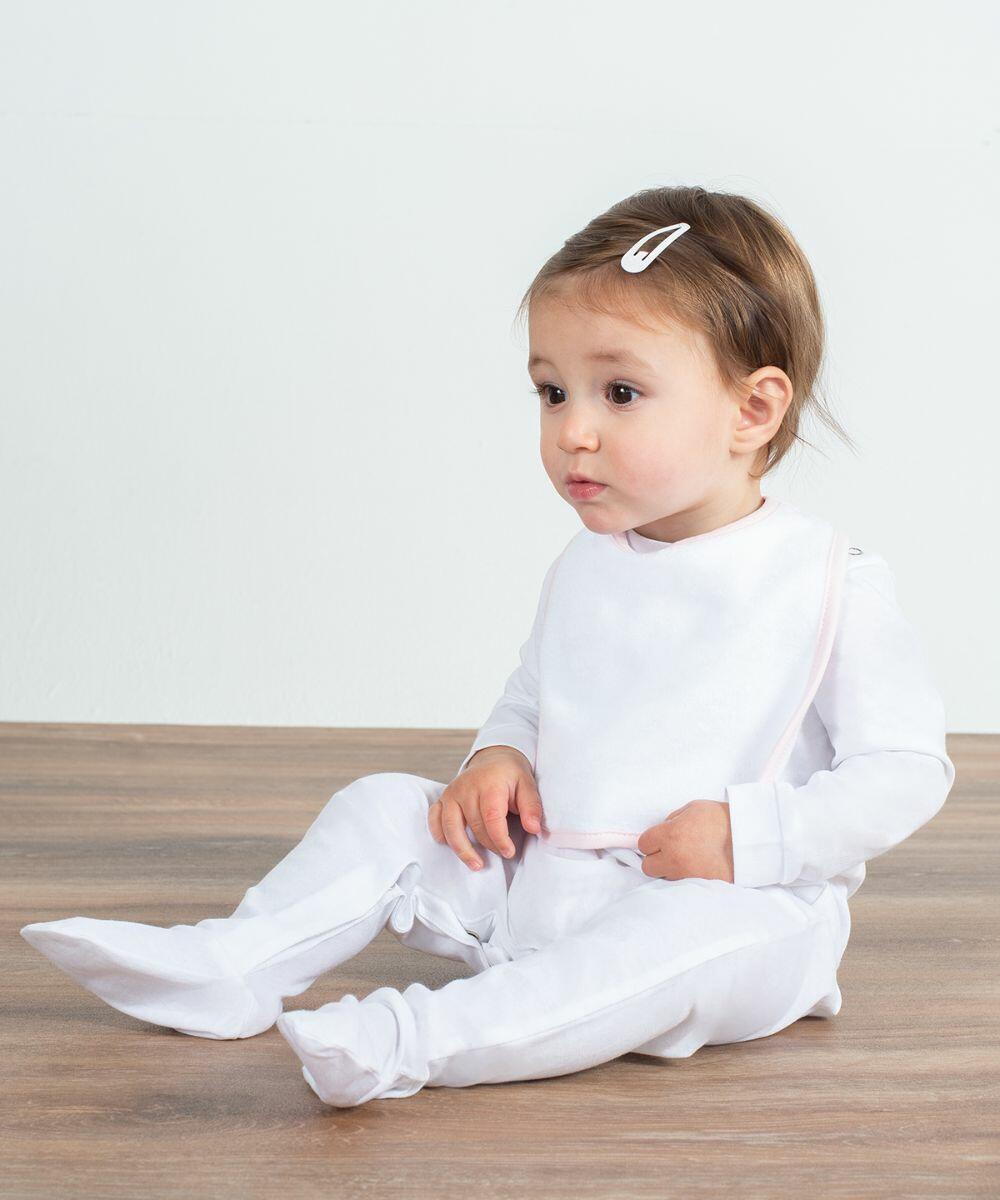 A baby wearing a white LW22T by Larkwood and a white bib, sitting on a wooden floor with a neutral expression. The baby has a small hairclip in their hair.