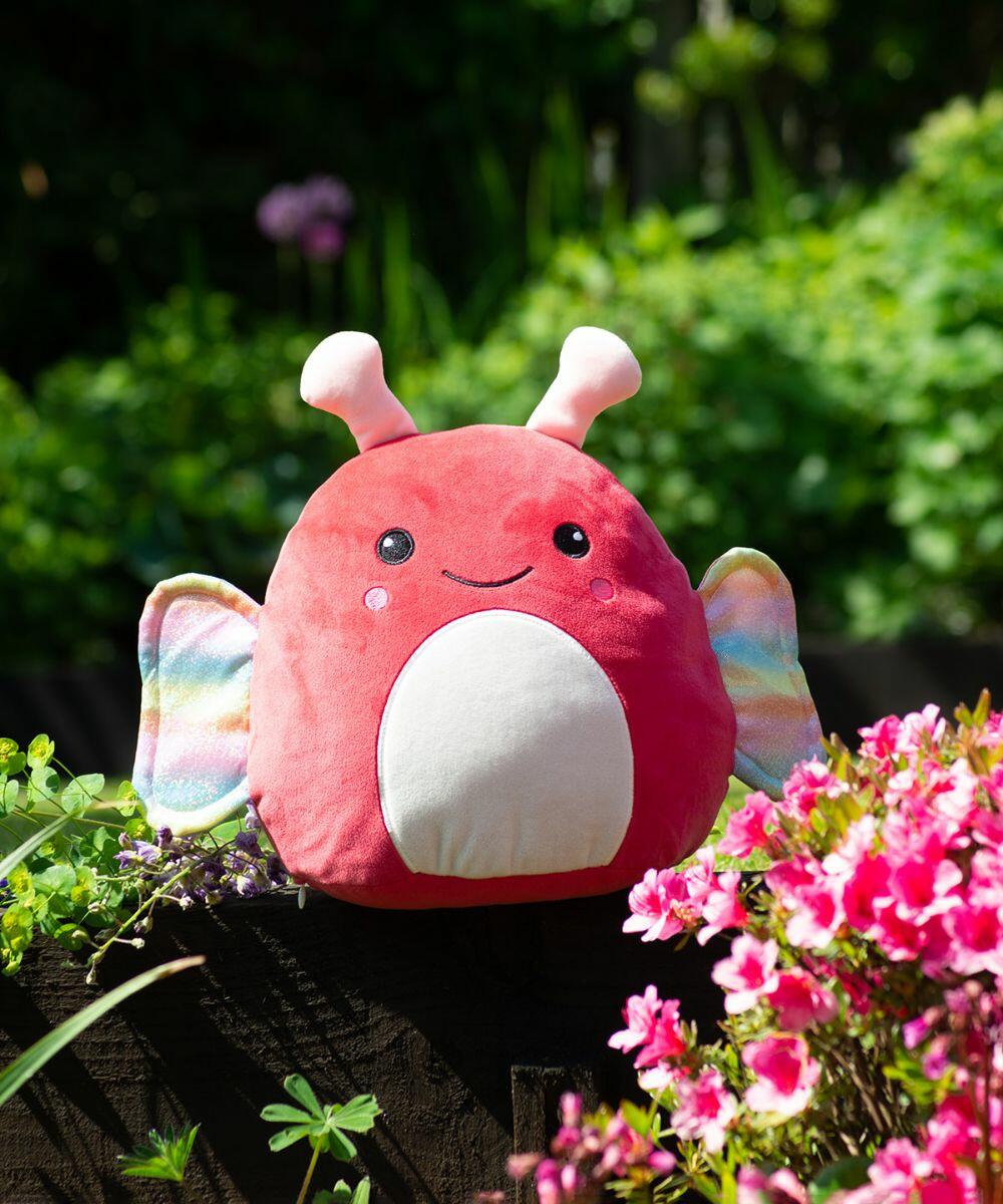 A red plush MM800 by Mumbles with a smiling face, antennae, and colorful wings sits among pink flowers and green plants in an outdoor setting.