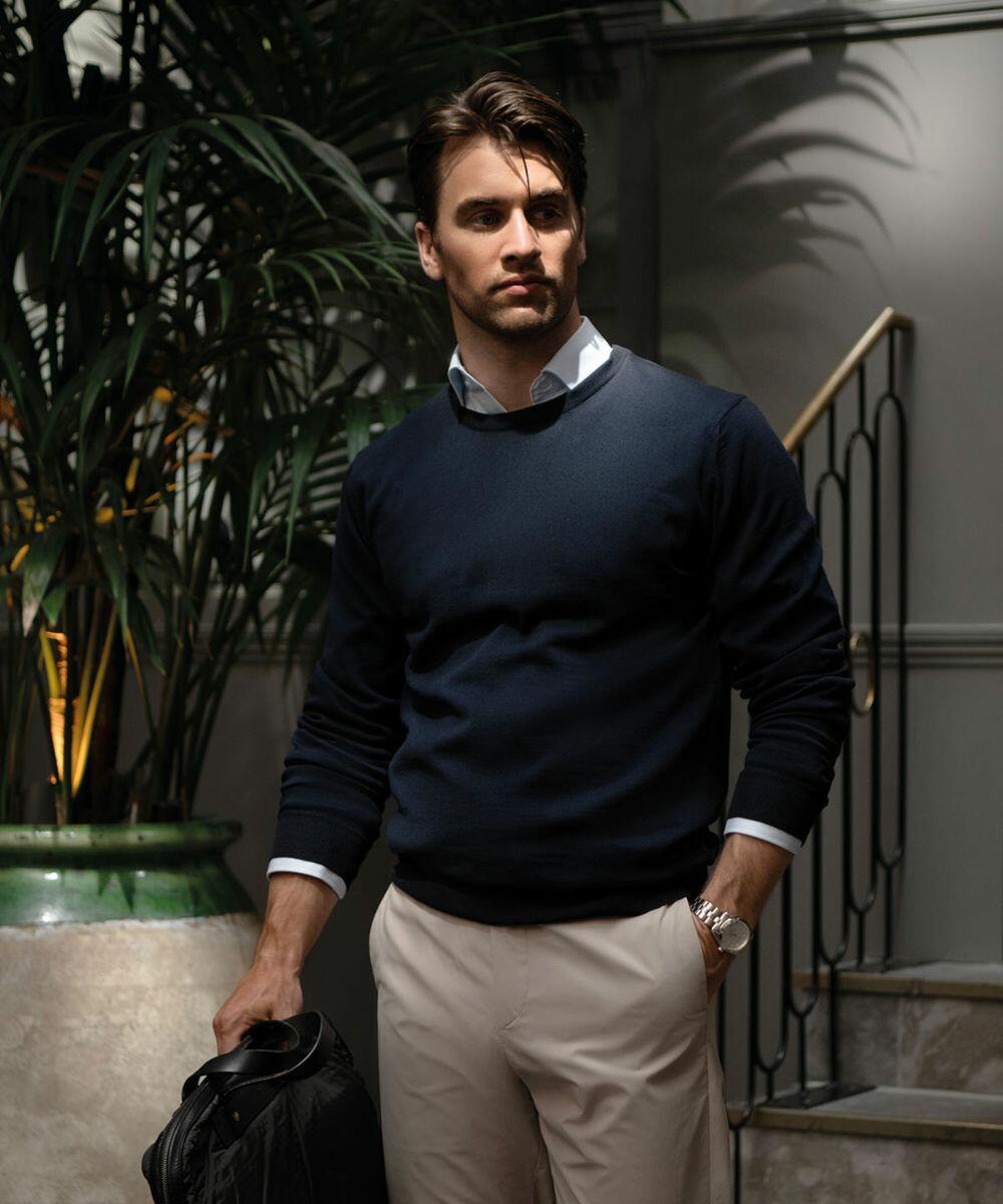 A man stands indoors near large potted plants, wearing a navy Nimbus N113M sweater, light collared shirt, and beige pants, holding a dark jacket in one hand.