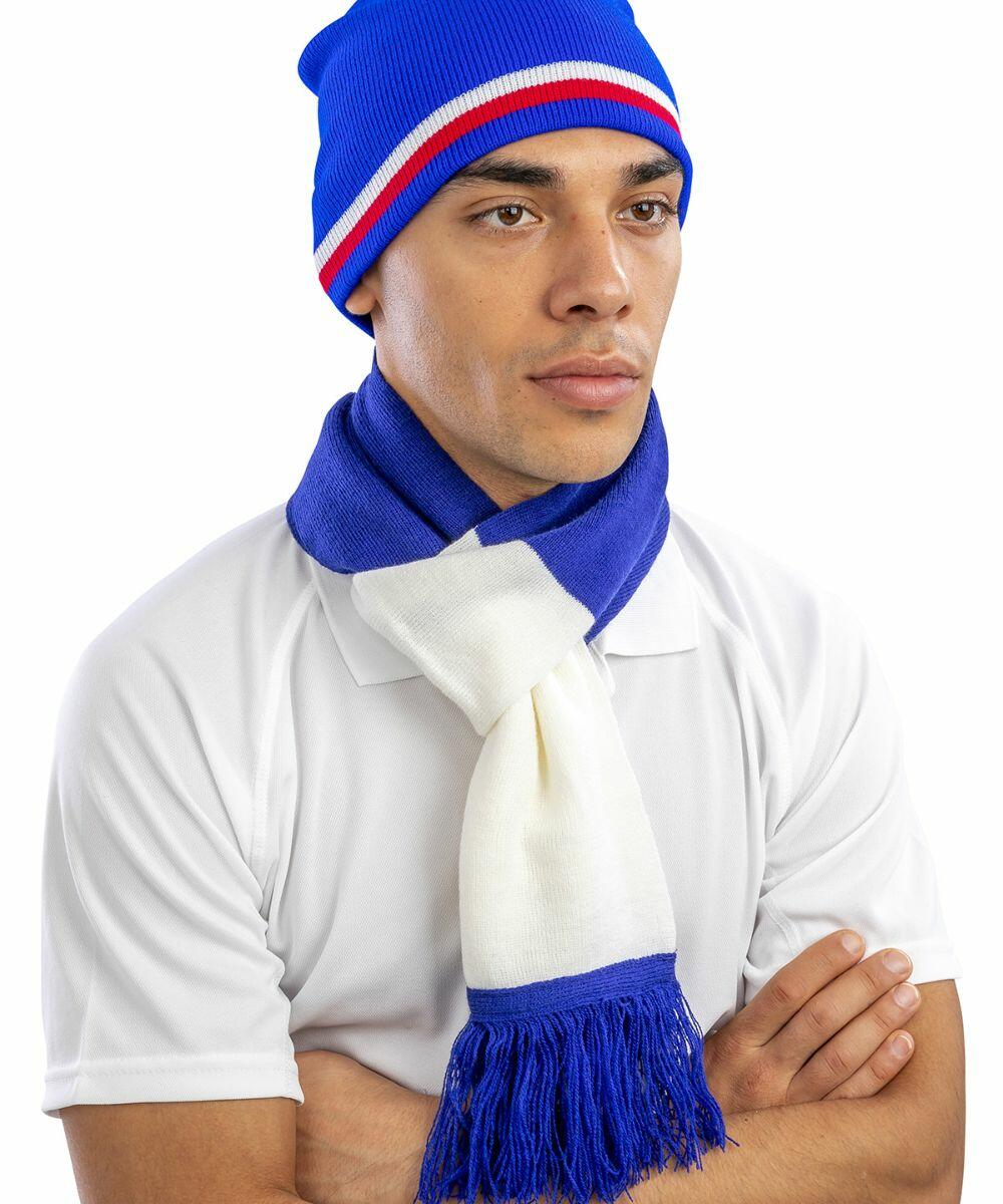 A person is wearing a blue, white, and red beanie and a blue and white scarf, both with fringe. They are dressed in a white shirt and have their arms crossed with a neutral facial expression, featuring the R146X from Result.
