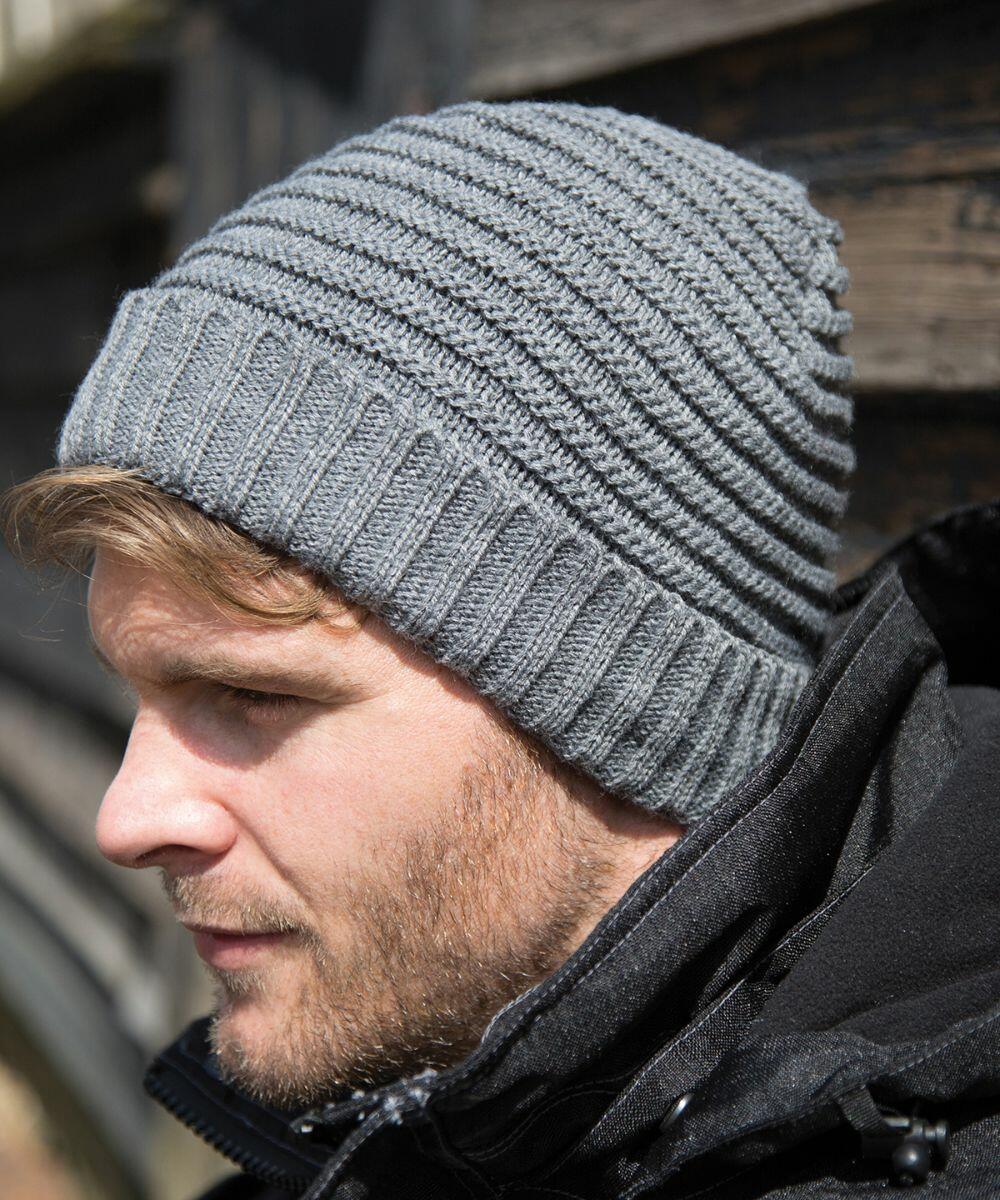 A person wearing a grey Result R376X knitted beanie and a black jacket, viewed from the side against a blurred wooden background.