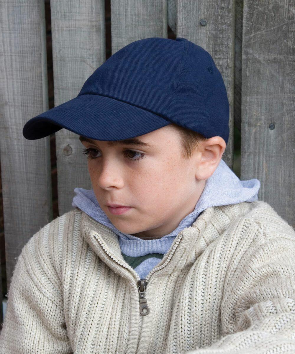 A young boy wearing a navy blue cap, a gray hoodie, and a beige knit sweater sits against a wooden fence looking to the side.