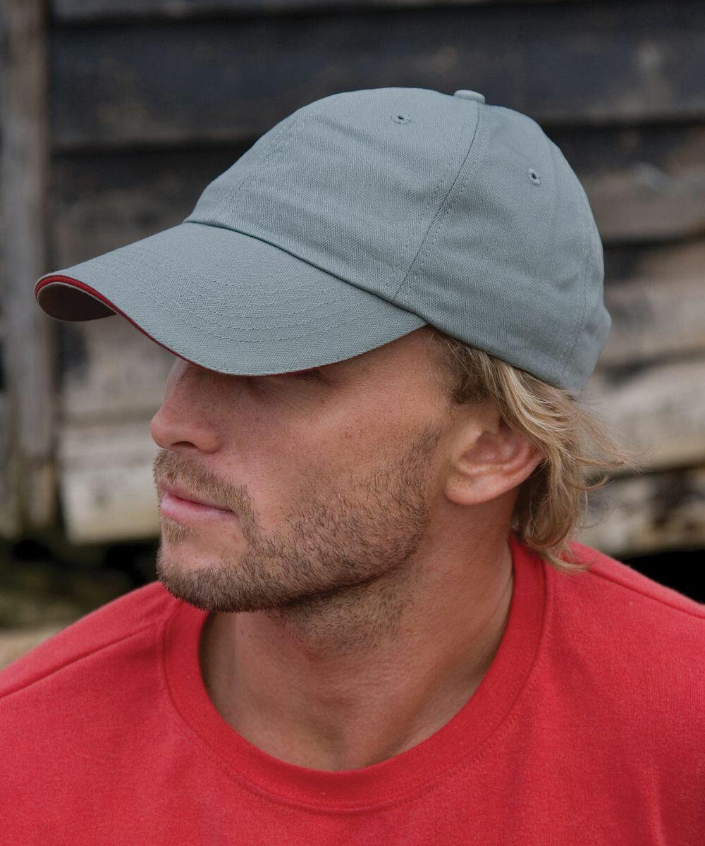 A man with a blonde beard is wearing a gray Result RC38X baseball cap and a red shirt, looking to his right. He is in front of a weathered wooden structure.