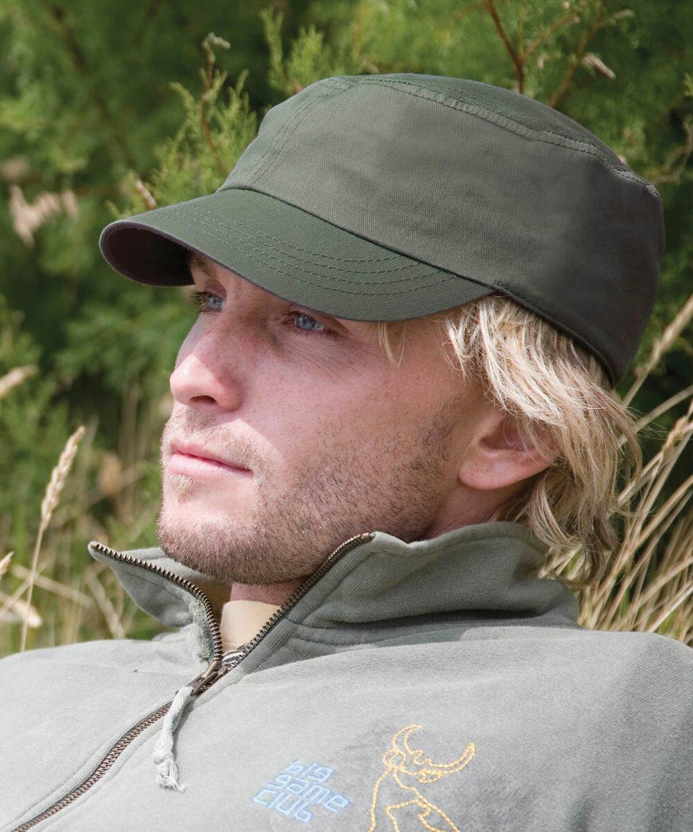 A person with blonde hair wearing an olive green cap and a grey Result RC70X jacket with an embroidered design, outdoors with greenery in the background.