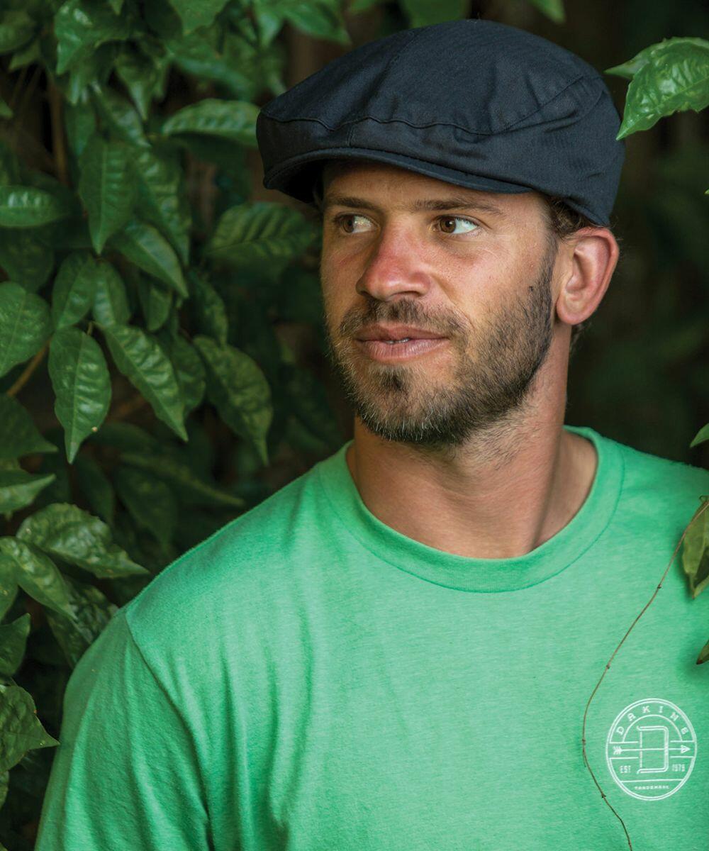 Man with a beard wearing a green RC77X T-shirt from Result and a flat cap, standing amidst green foliage.