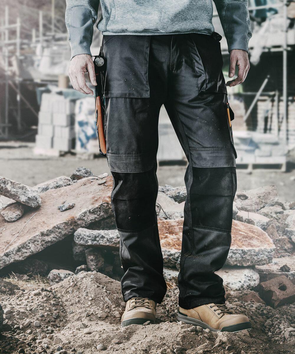 A person wearing Scruffs SH056 protective pants and work boots stands in a construction site with rubble and unfinished structures in the background.