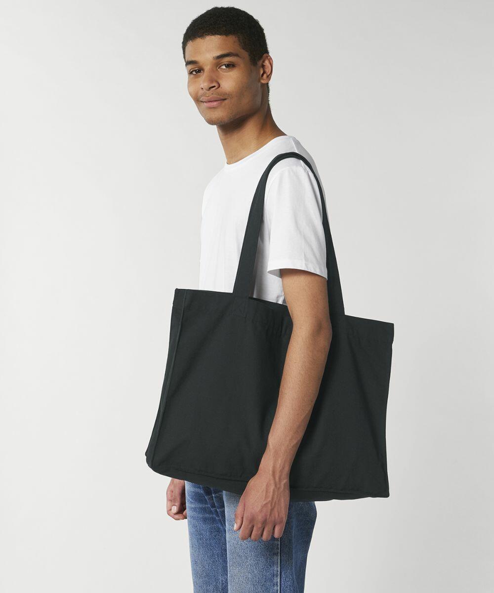 A person stands sideways wearing a white t-shirt and blue jeans, carrying a large black Stanley/Stella SX062 tote bag on their shoulder.