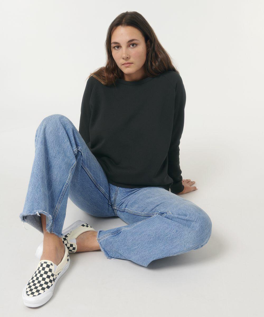 A person with long hair in a black Stanley/Stella SX083 sweatshirt and blue jeans sits on the floor with legs crossed and hand resting behind them. They are wearing checkered slip-on shoes.