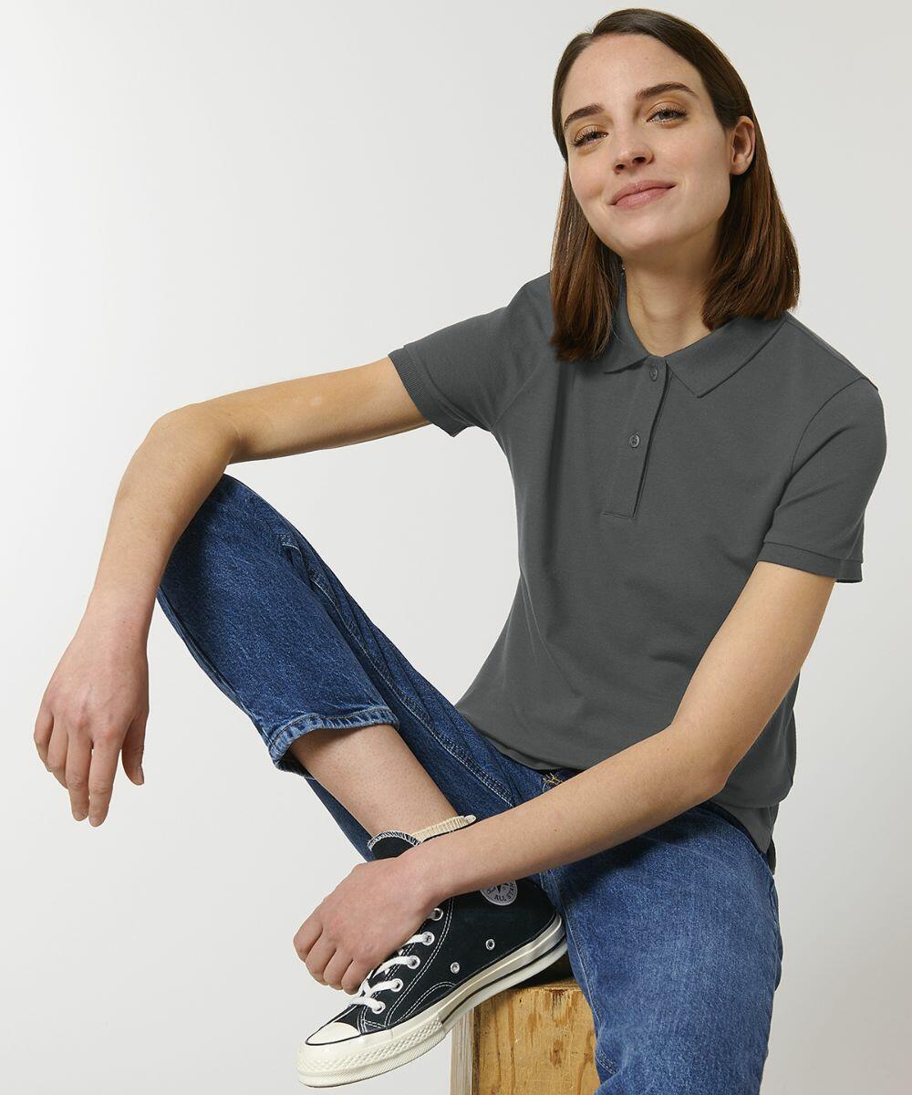 A person with shoulder-length hair wearing a gray Stanley/Stella SX131, blue jeans, and black sneakers is sitting on a wooden box against a plain background.