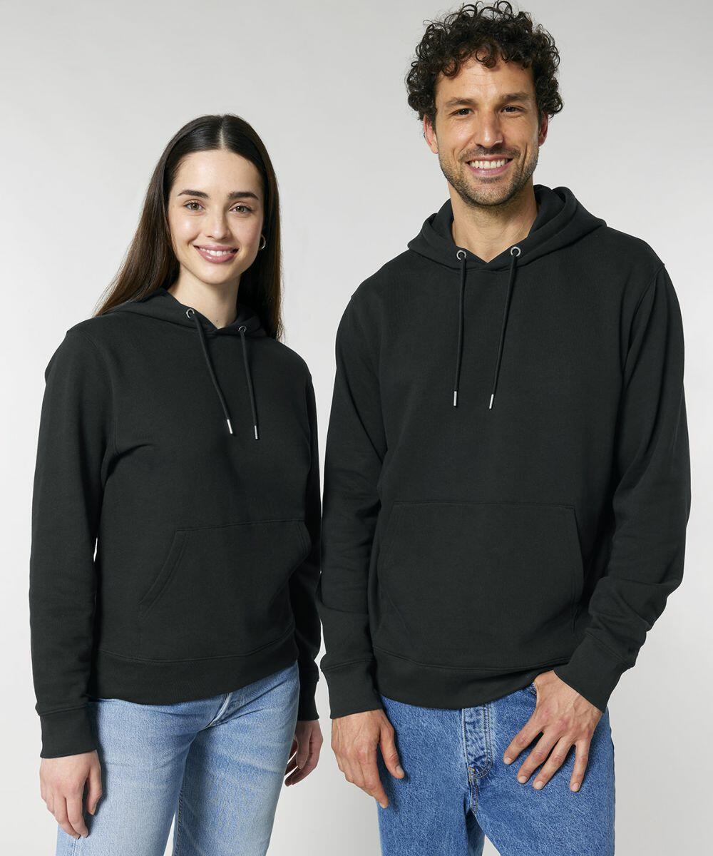 Two people, one with long dark hair and one with short curly hair and a beard, are standing and smiling. Both are wearing Stanley/Stella SX705 hoodies and blue jeans against a plain white background.