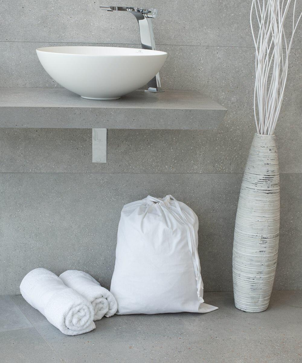 A sleek bathroom with a vessel sink, a neatly rolled pair of white Towel City TC063 towels, a white drawstring laundry bag, and a tall decorative vase with white branches placed on a gray concrete floor.