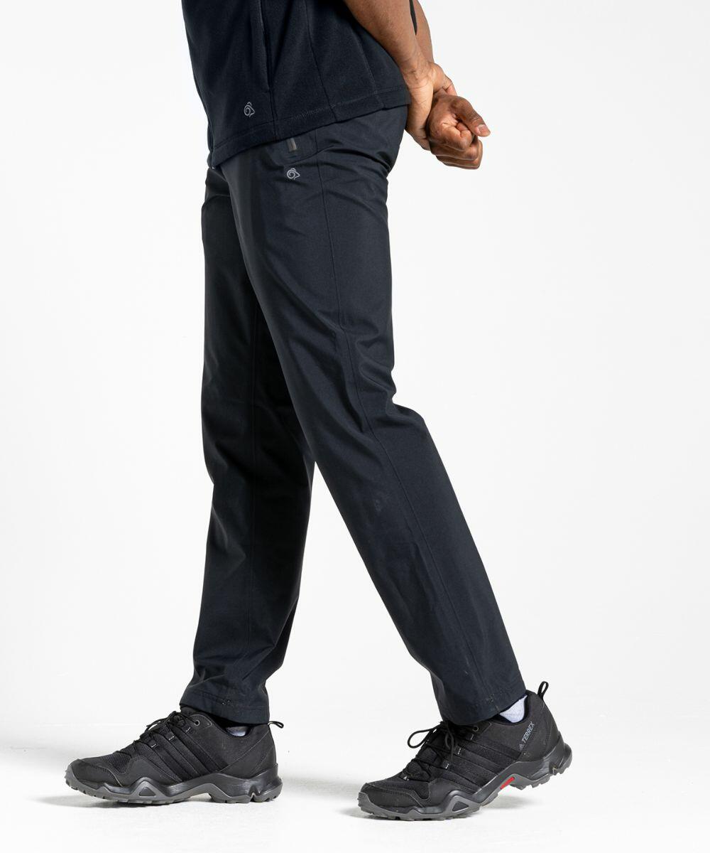 A person wearing black pants and black Craghoppers CR237 sneakers stands with hands clasped behind their back, facing slightly to the right. The background is plain white.