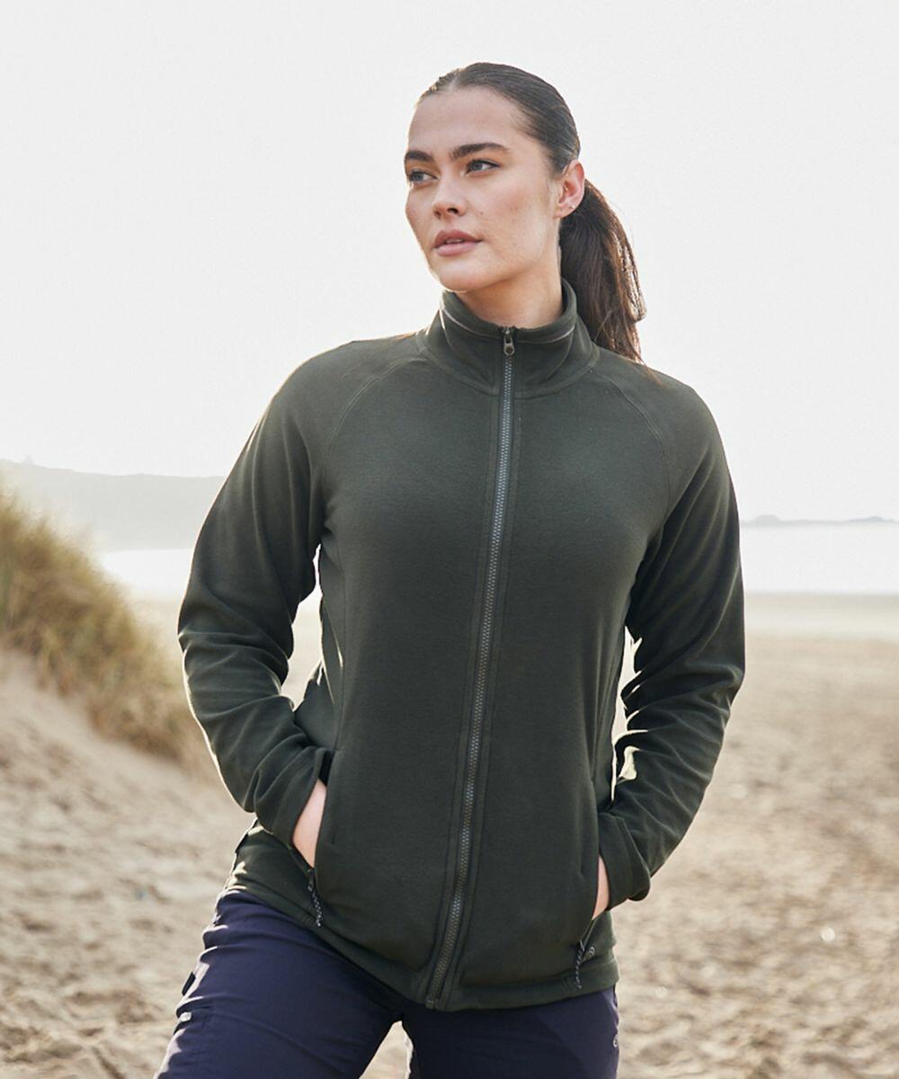 A person stands on a beach wearing a dark green Craghoppers CR316 zip-up jacket and dark pants, with hands in pockets and looking into the distance.