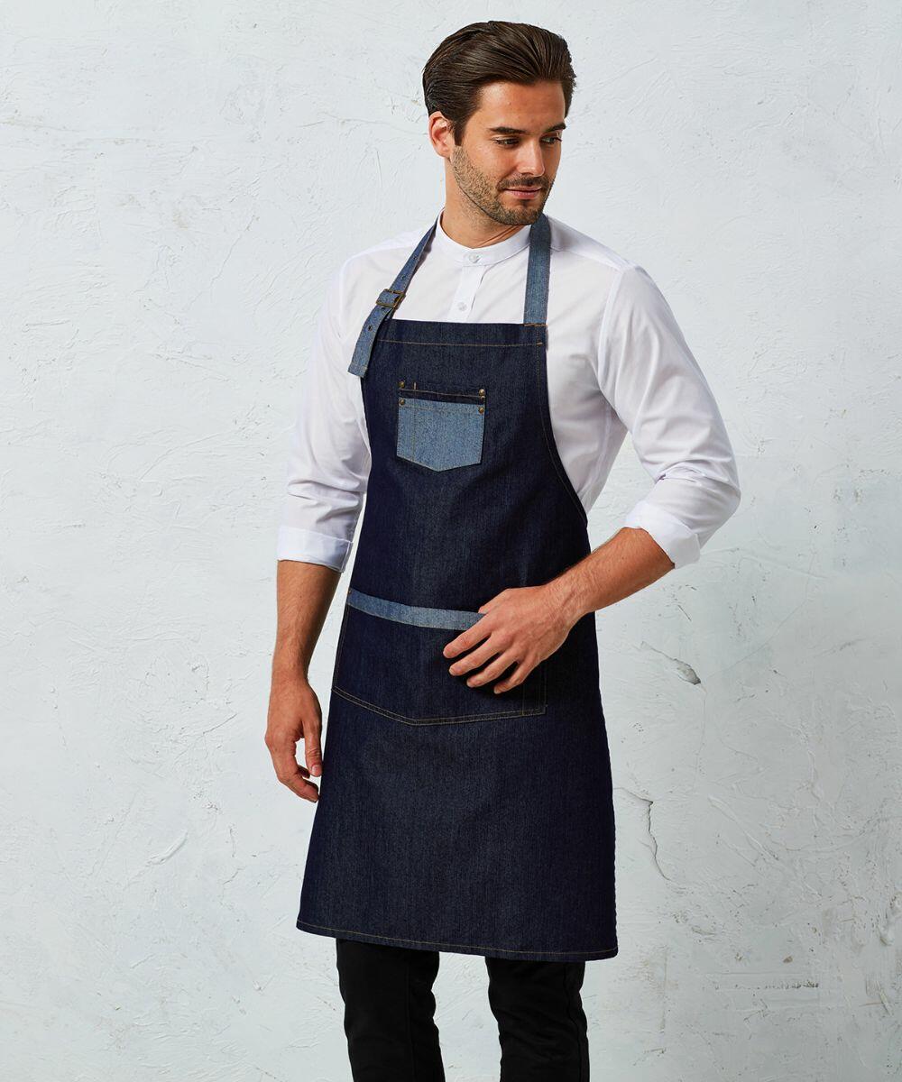 A man wearing a white shirt and a Premier PR127 dark denim apron with a front pocket stands against a light-colored wall.