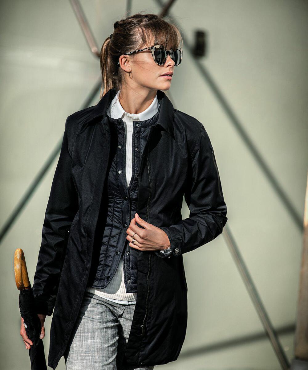 A person wearing a black coat, sunglasses, and plaid pants holds a Nimbus NB51F umbrella while standing outdoors against a modern architectural background.
