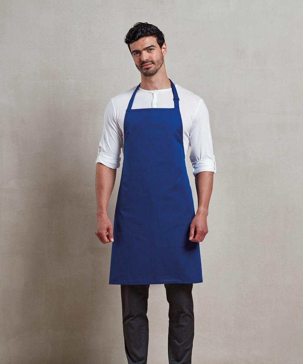 A man in a white shirt and dark pants is wearing a blue Premier PR996 apron, standing against a plain background.