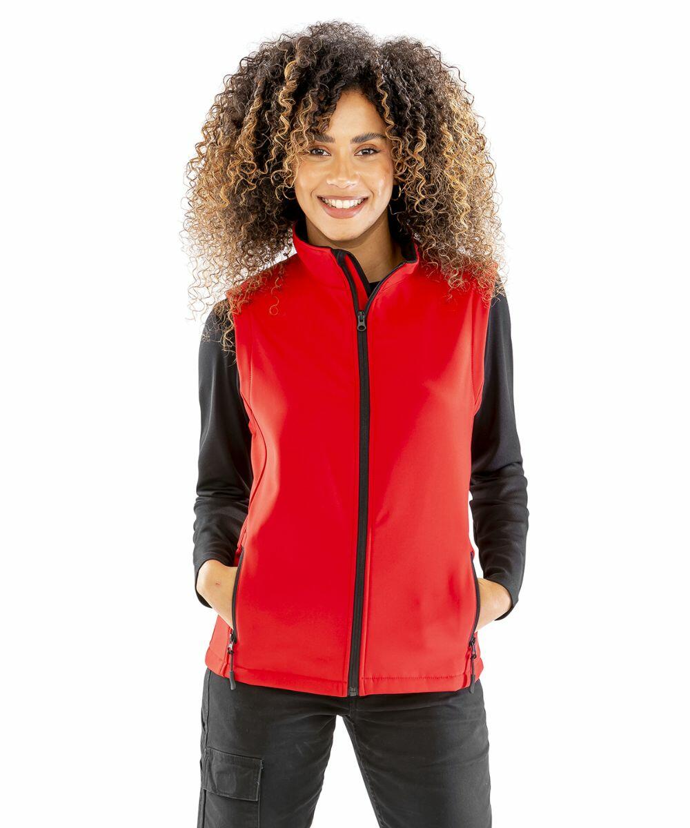 A person with curly hair is smiling, wearing a red Result R232F zip-up vest over a black long-sleeve shirt, and dark pants, standing with hands in pockets.