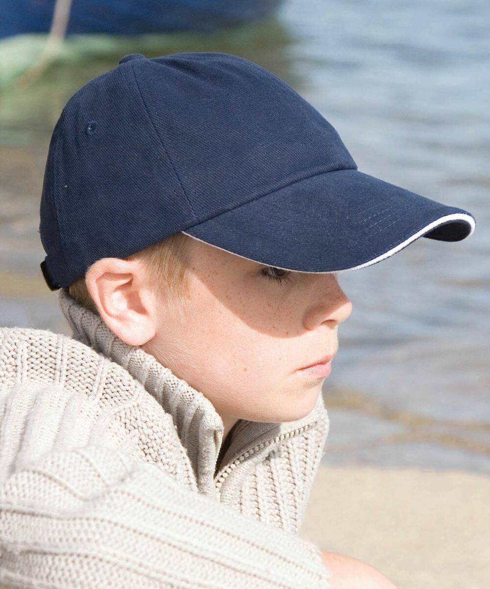 A child with a navy baseball cap sits by the water, wearing a Result R24JP knit sweater, and looking into the distance.