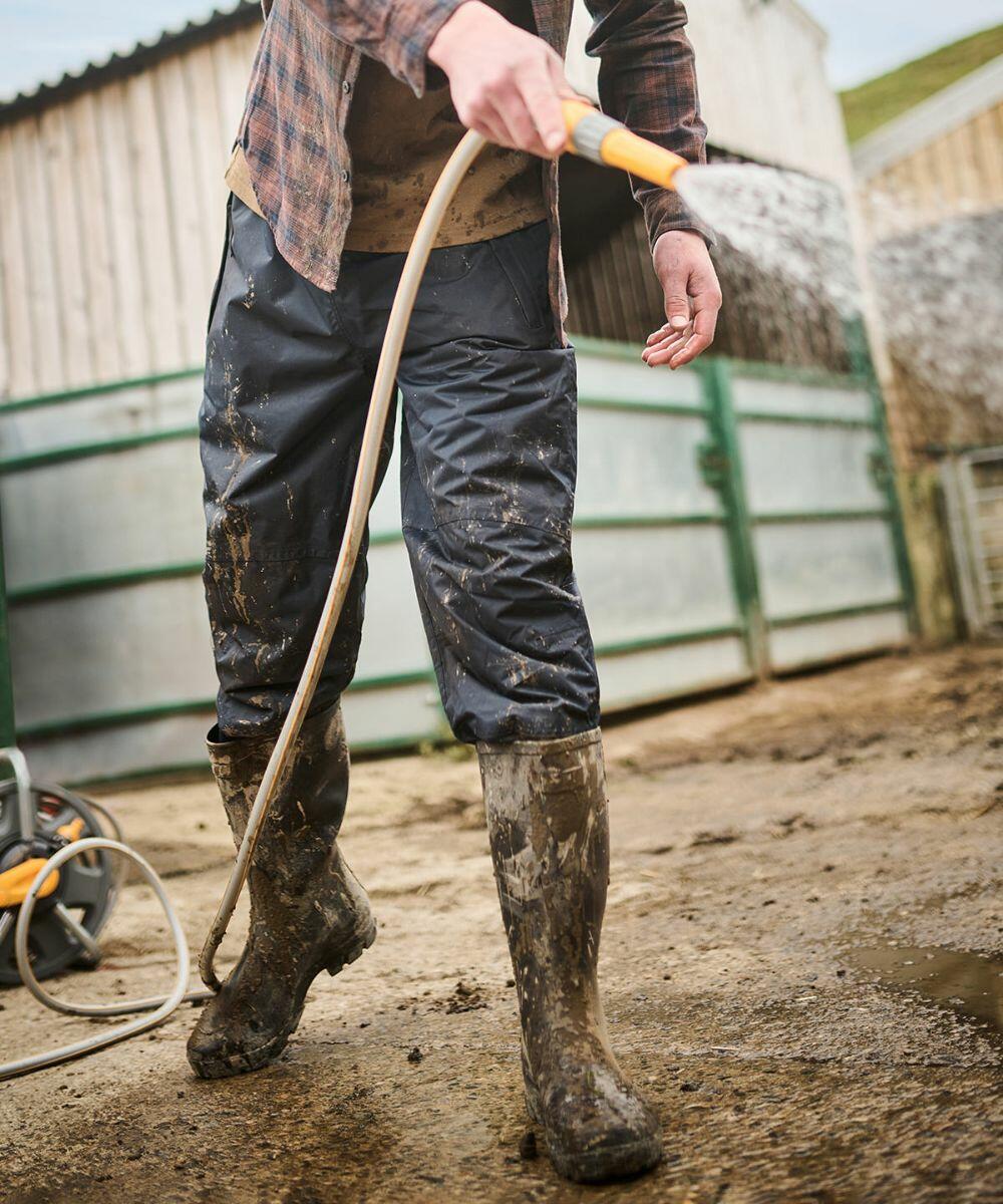A person wearing muddy boots and pants is using a Regatta RG030 hose to spray water, likely cleaning an outdoor area near a wooden structure and metal fence.