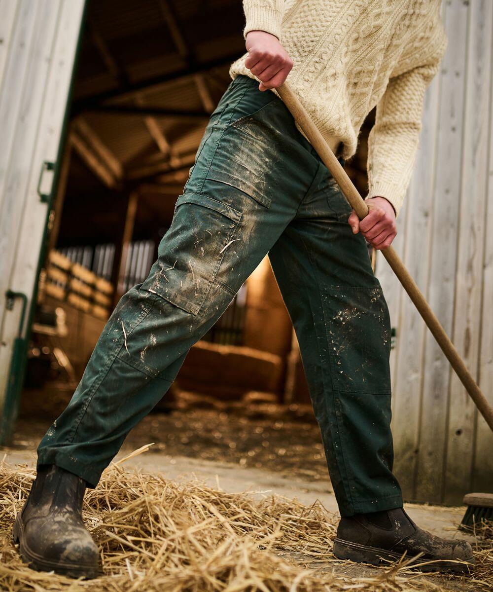 Person wearing a Regatta RG232 sweater, green pants, and boots, holding a rake and working with hay inside a barn.