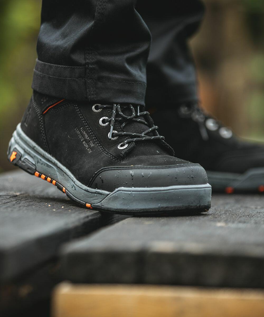 Close-up of a person wearing SH083 work boots by Scruffs with grey soles and orange accents, standing outdoors on a wooden surface.