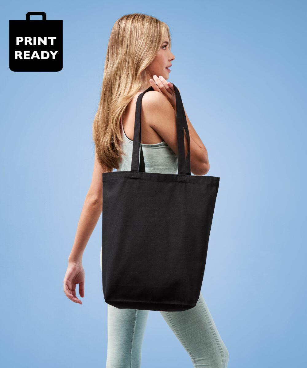 A person in a light green outfit is carrying a Nutshell RL660 black tote bag over their shoulder. The background is blue, and there's a "Print Ready" logo in the top left corner.