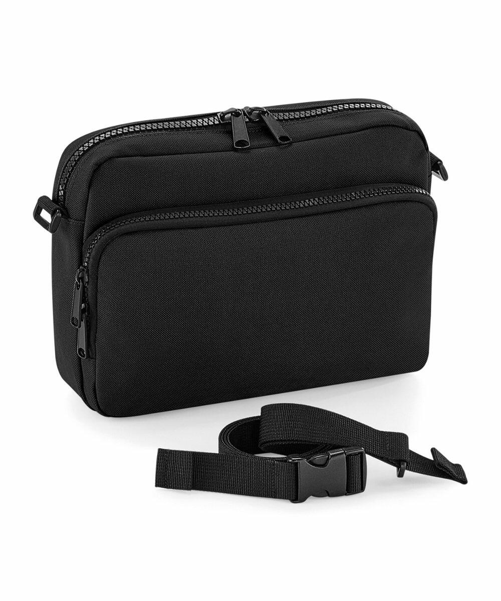 A black, rectangular BG242 shoulder bag by Bagbase with two zippered compartments and a detachable, adjustable strap, placed in front of the bag. The bag has a minimalist design with smooth fabric and sturdy zippers.