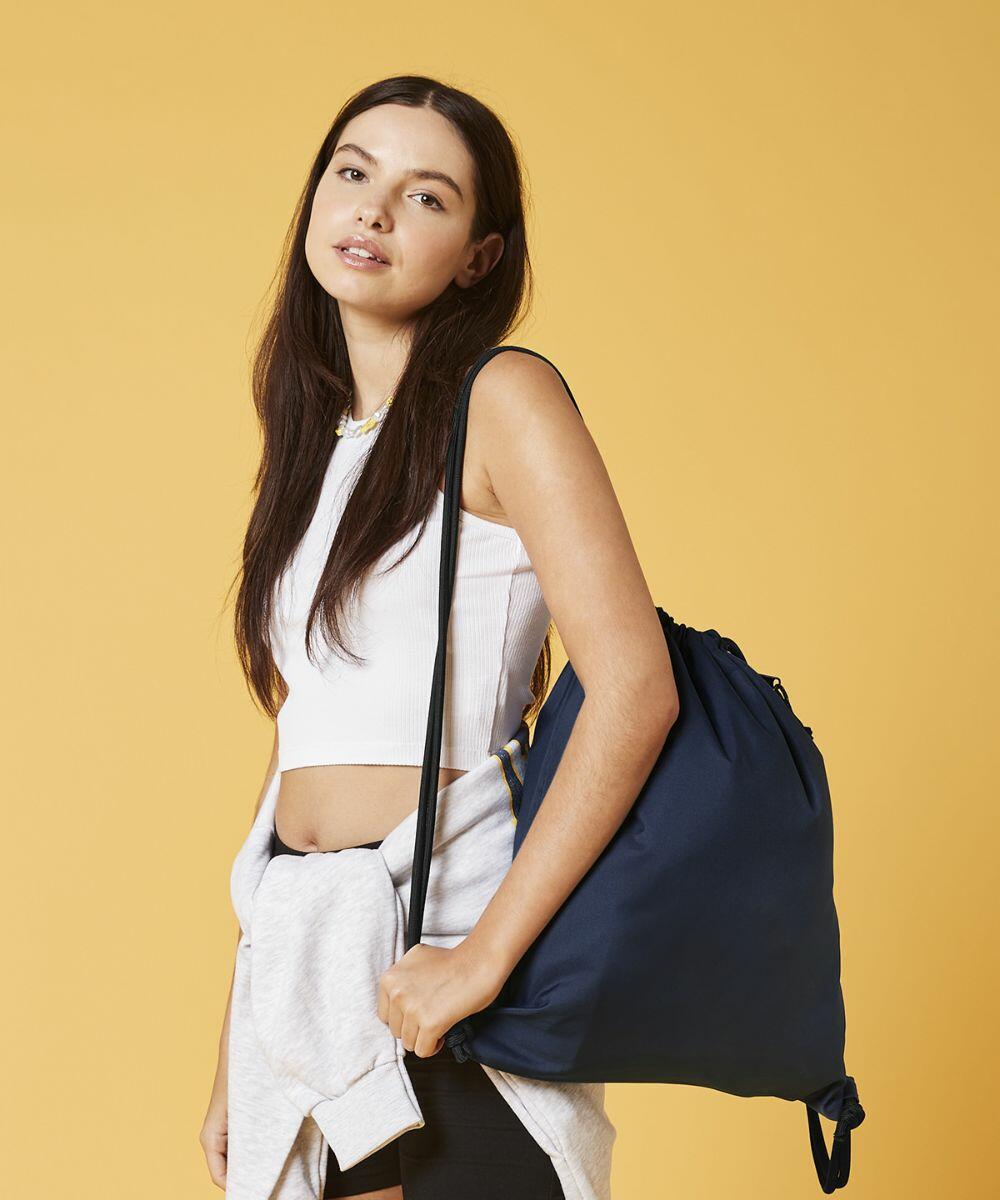 A young woman with long, dark hair stands against a yellow background. She is wearing a white cropped tank top and has a light gray sweatshirt tied around her waist. She carries a navy blue Bagbase BG281 over her shoulder and looks at the camera with a slight smile.