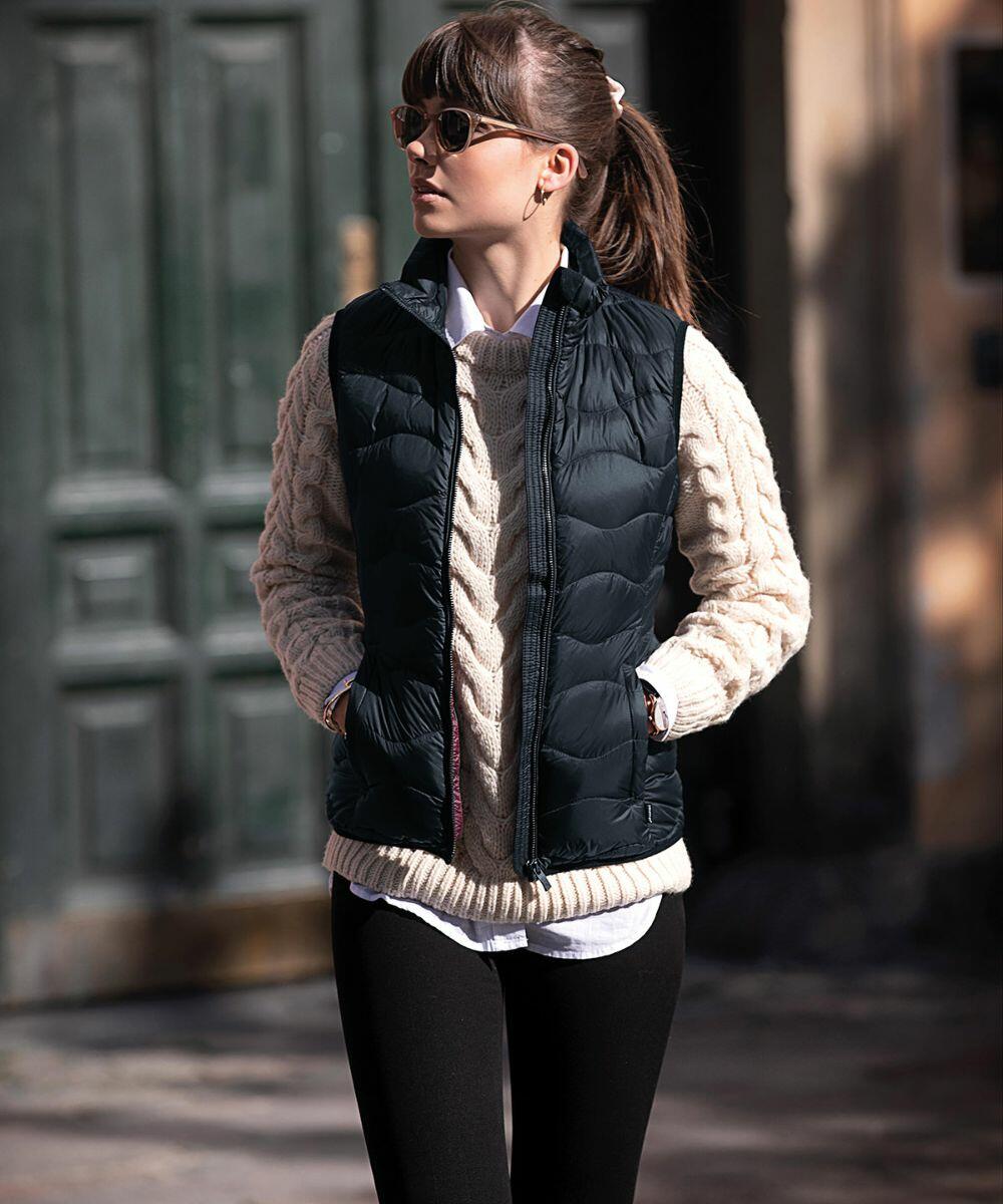 Person wearing a Nimbus NB79F dark quilted vest over a cable-knit sweater and black pants, standing outdoors with hands in vest pockets, looking to the side.