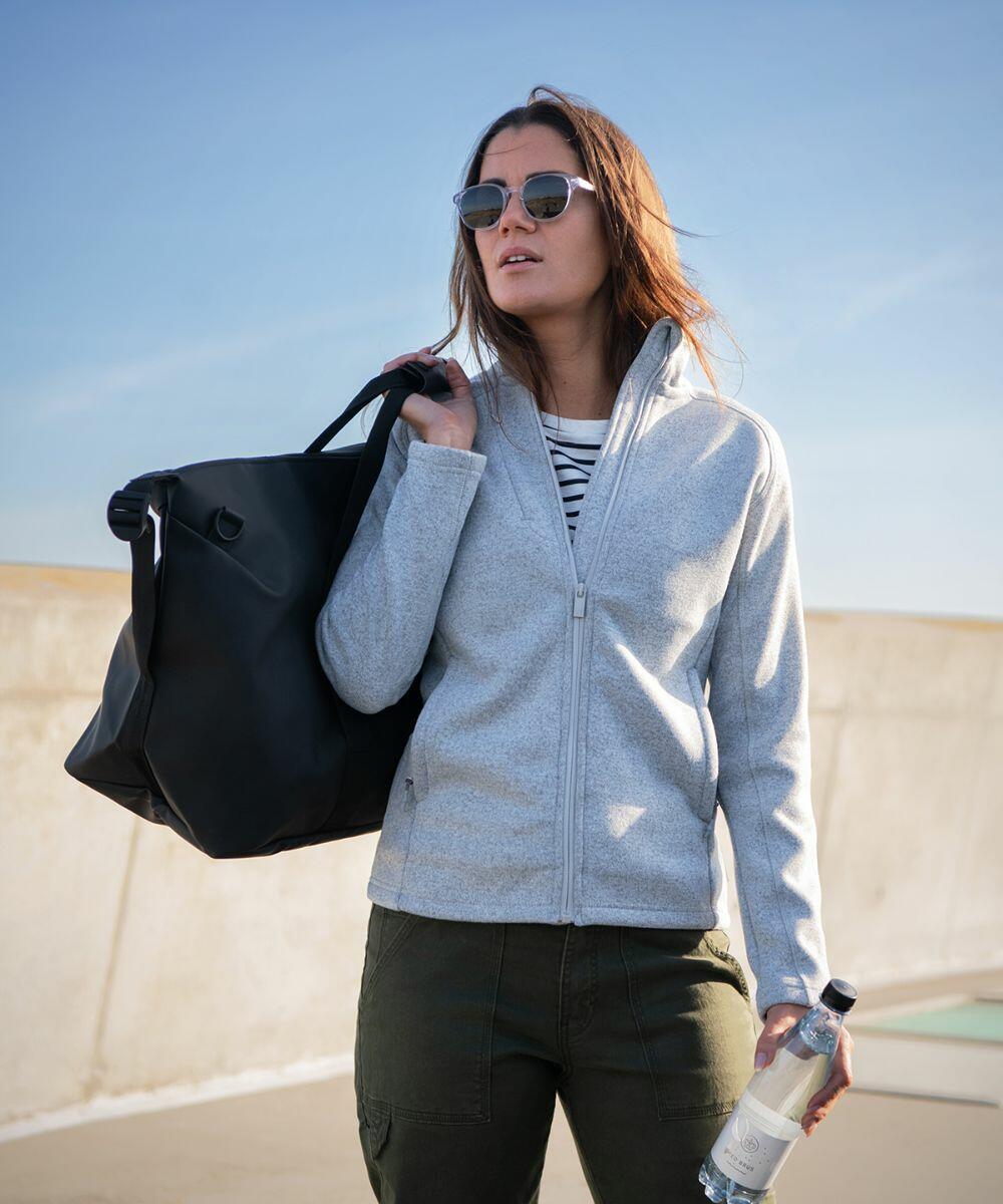 A woman wearing sunglasses, a grey zip-up jacket, and green pants holds a large Nimbus NP08F on her shoulder and a plastic water bottle in her left hand. She stands outdoors with a blue sky background.