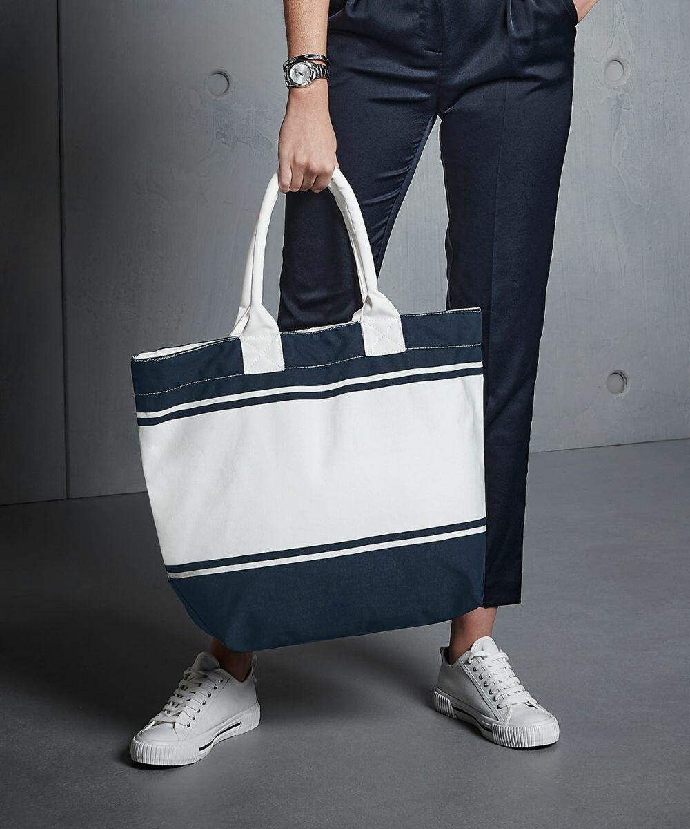 Person holding a large white Quadra QD026 tote bag with dark blue stripes, dressed in dark blue trousers, white sneakers, and a silver watch. Background is a gray, modern, industrial-style wall.