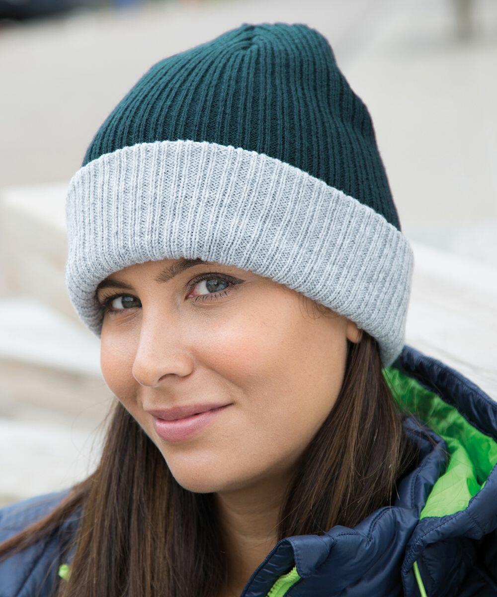 A person wearing a dark green and light gray beanie with a slight smile, dressed in a Result R378X jacket with a green inner lining, is shown in an outdoor setting.