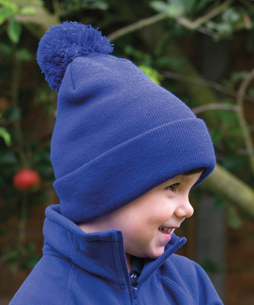 A child in a blue Result RC28J hat with a pom-pom and a matching blue jacket smiles, while standing outside near some foliage.