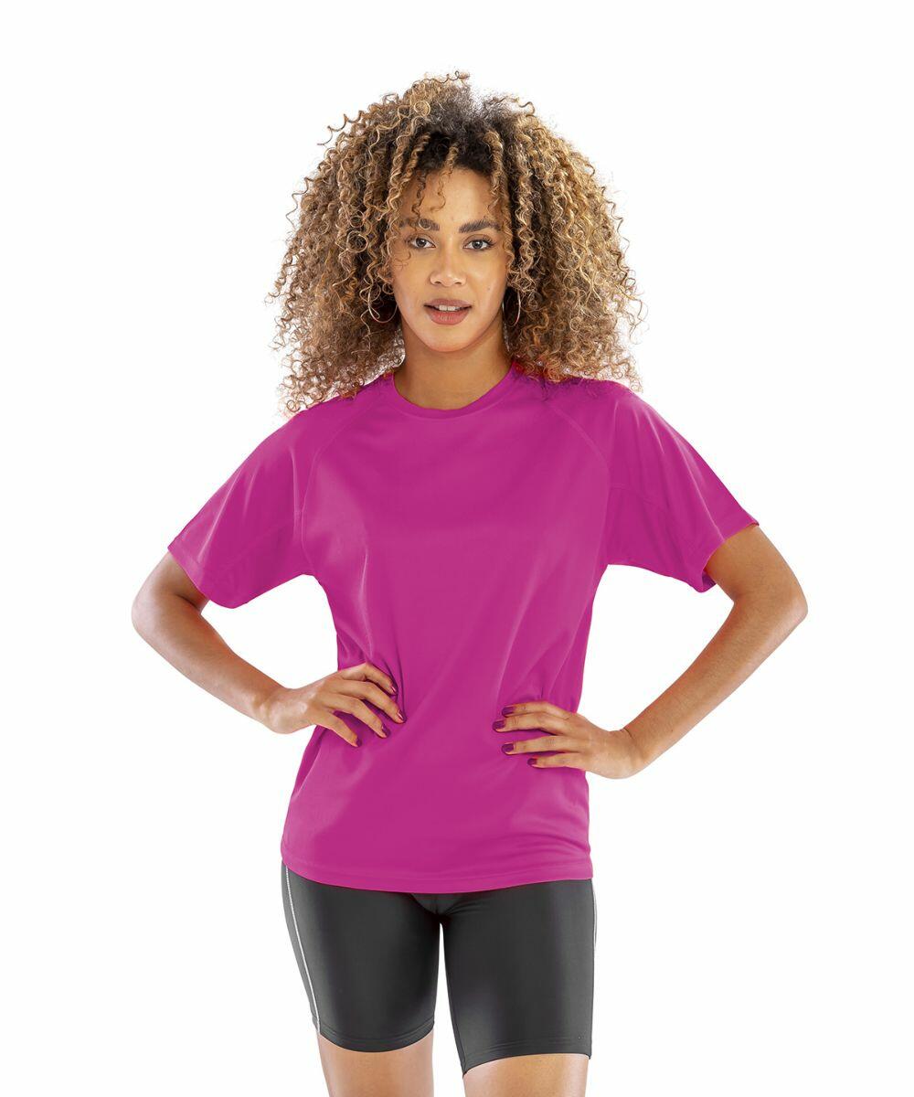 A person with curly hair stands confidently wearing a Spiro S287X bright pink T-shirt and black shorts, against a plain white background.
