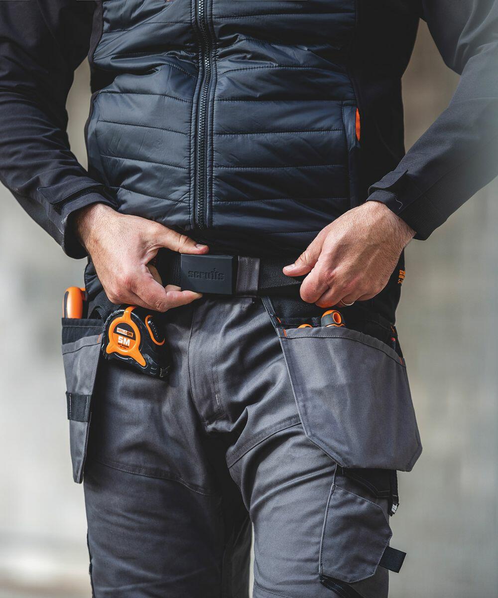 Person wearing a black jacket and gray work pants, adjusting a Scruffs SH031 tool belt with various tools and a tape measure.