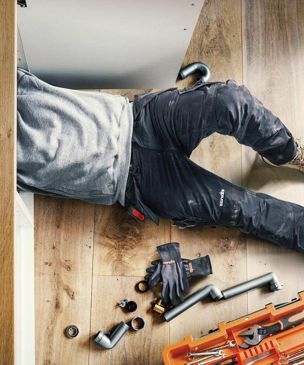 Person lying on the floor, partially under a cabinet, working on plumbing. Tools, gloves, and pipe fittings scattered around. Scruffs SH071 orange toolbox and wooden floor visible.