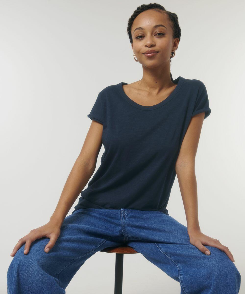 Person wearing a navy blue t-shirt and blue jeans, seated on a stool against a plain background, looking at the camera with a neutral expression while showcasing the SX084 from Stanley/Stella.