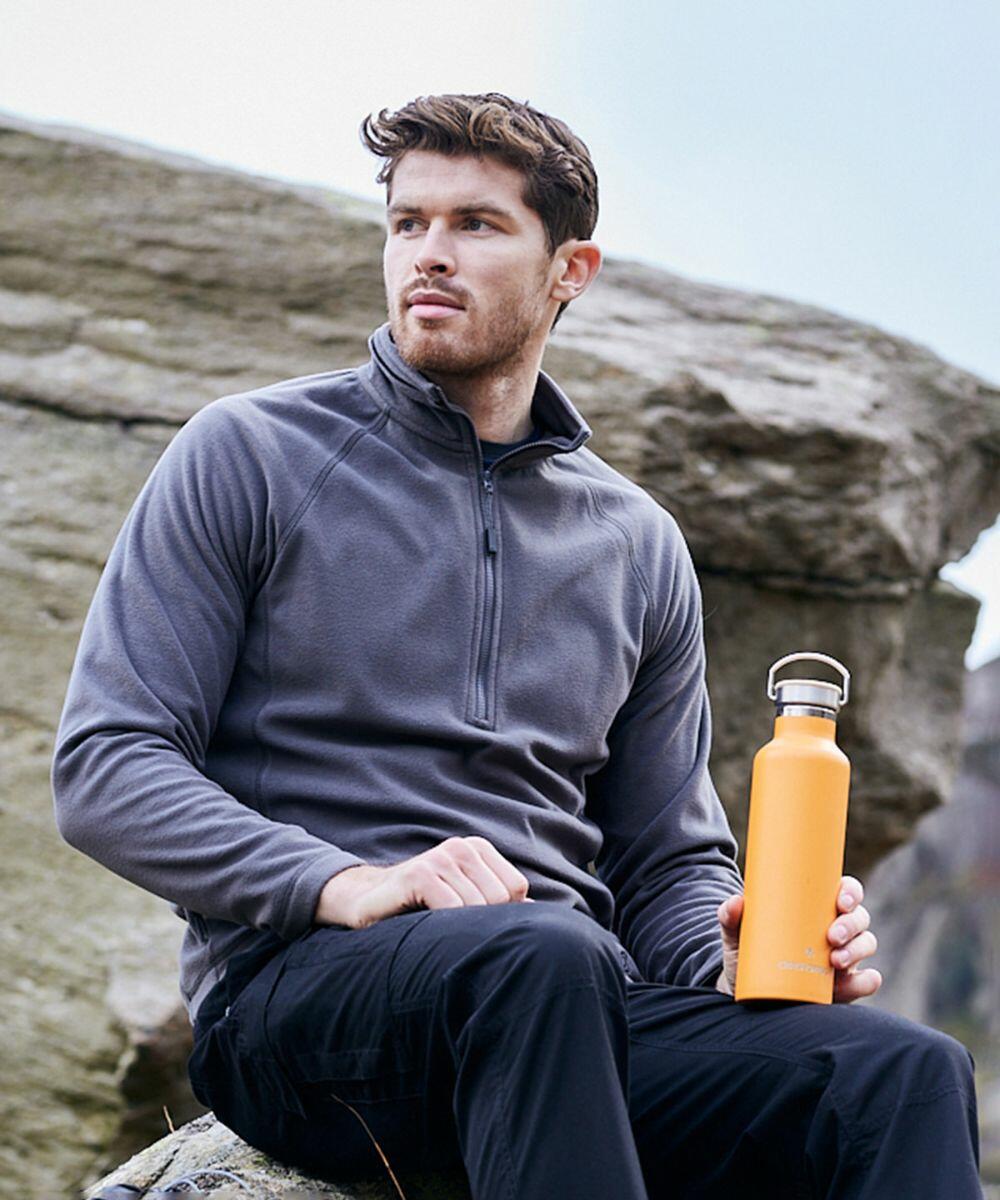 A person wearing a gray jacket sits on a rock, holding a Craghoppers CR317 water bottle.
