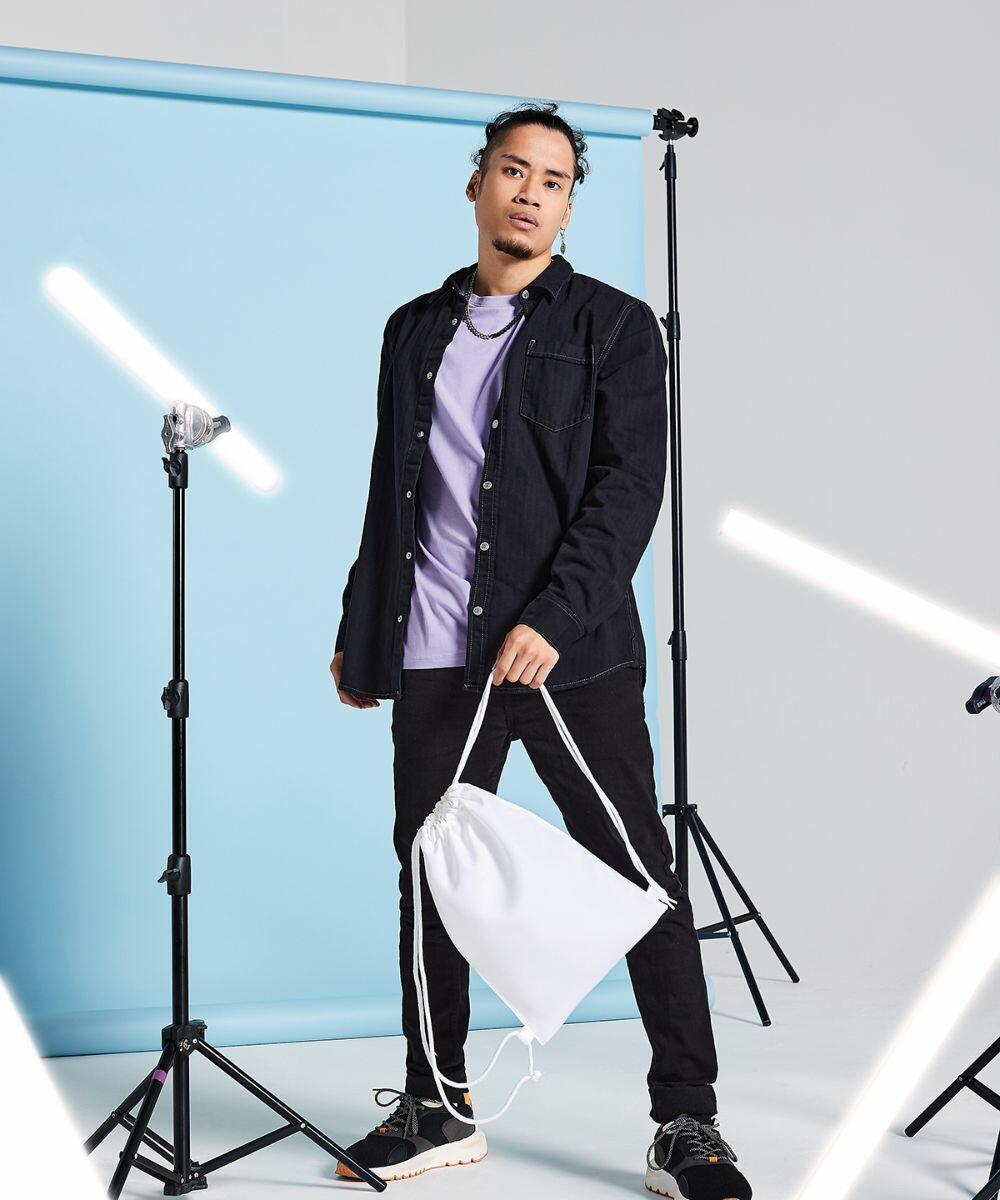 A person stands in a studio with a light blue backdrop, holding a white Bagbase BG910 drawstring bag. They wear a black jacket, lavender shirt, black pants, and white sneakers. Lighting equipment is visible around them.