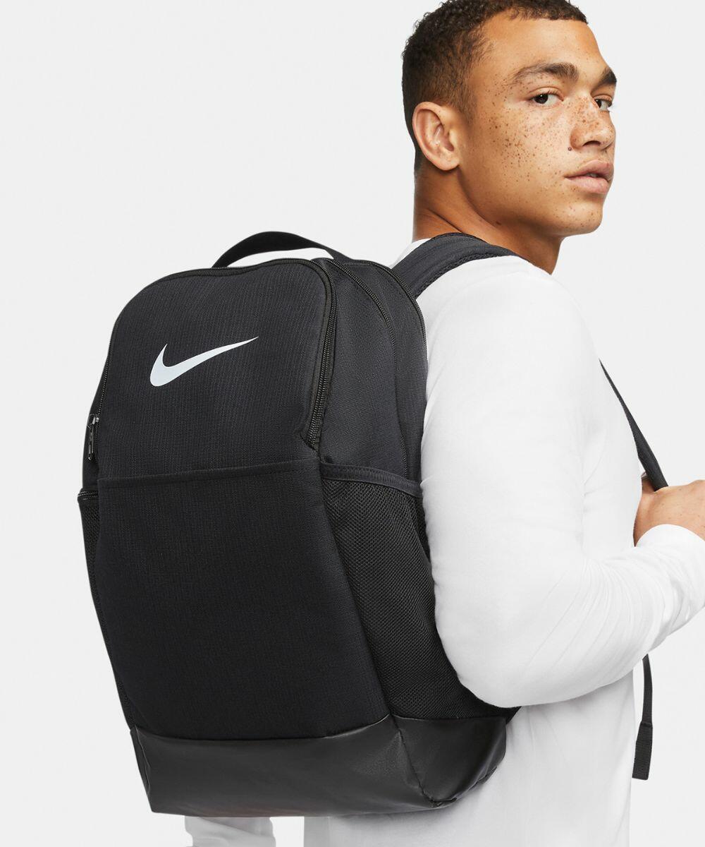 Person wearing a white shirt and carrying a black Nike NK351 backpack with a logo on the front.