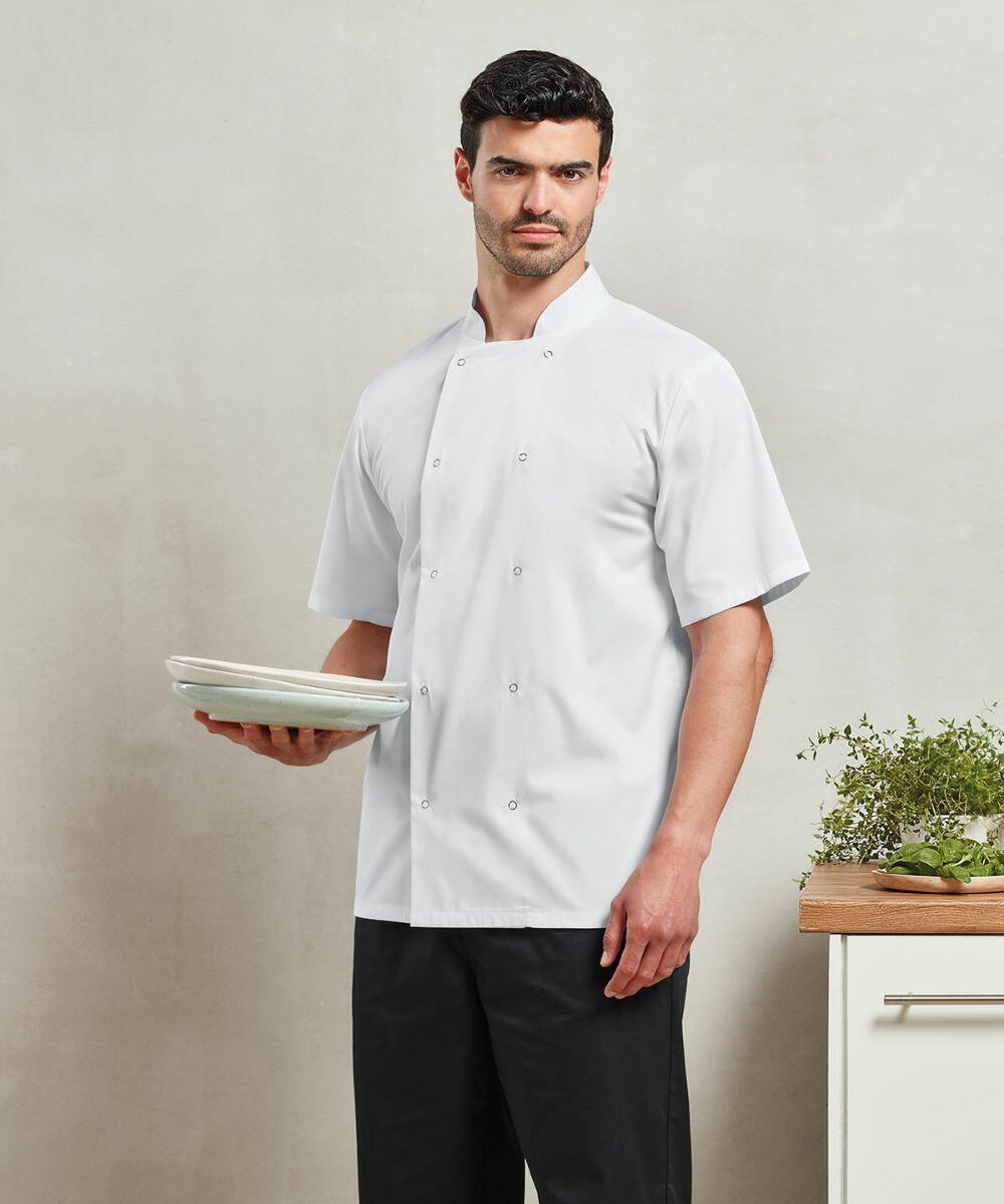 A man in a white chef's coat and black pants stands holding a stack of Premier PR664 plates. He is in a kitchen setting with a countertop and herbs nearby.
