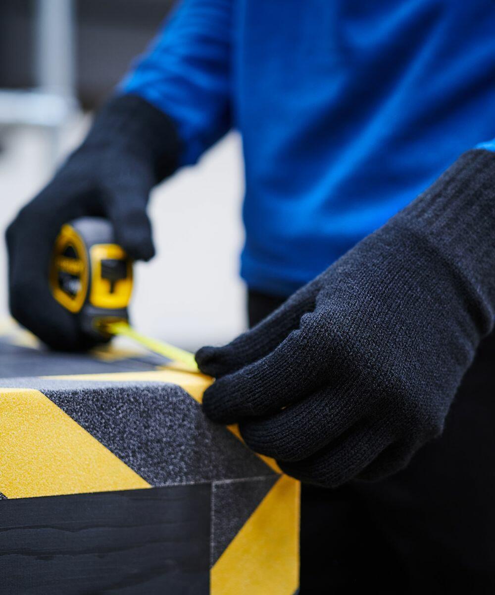 A person wearing black gloves uses a Regatta RG277 tape measure on a box with yellow and black striped tape.