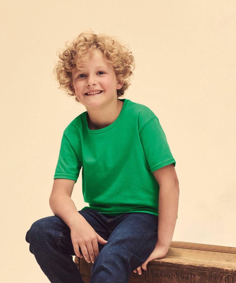 A young boy with curly hair is sitting on a wooden crate, wearing a green Fruit of the Loom SS023 T-shirt and jeans, smiling at the camera.